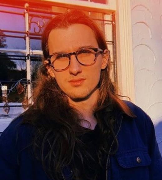 Person with long brown hair and round glasses in warm sunset light by a window