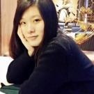 A woman with long black hair rests her chin on her hand, sitting in a cozy setting.